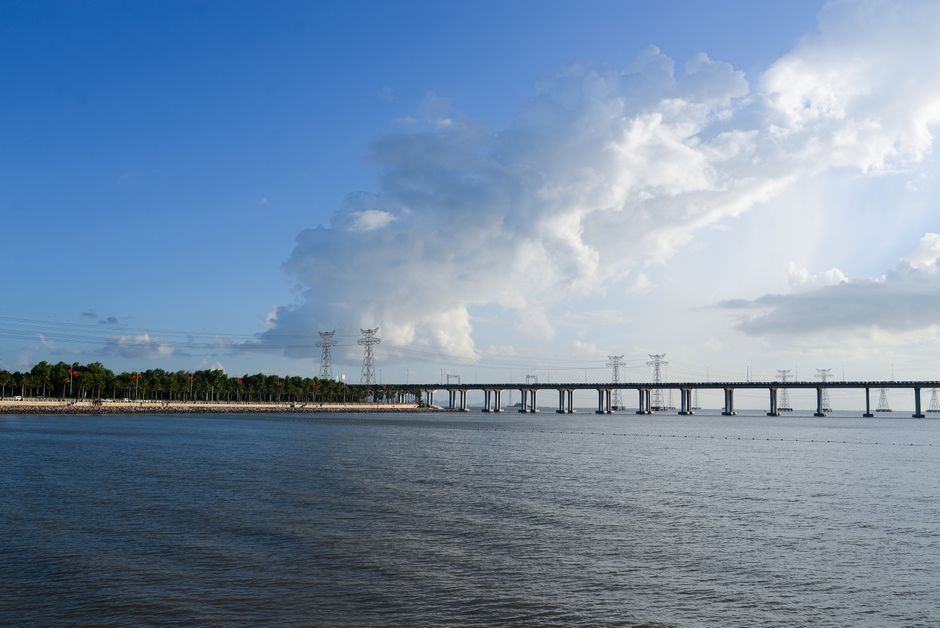 Photograph of a large body of water with a bridge extending across the horizon, supported by multiple concrete piers, under a partly cloudy sky with sunlight breaking through the clouds. In the background, a line of trees runs parallel to the shoreline. The scene captures a calm, expansive waterway with the bridge used for vehicular transport. No furniture, boxes, or moving equipment are visible in the image, and it does not depict any ongoing removals or packing activities. This landscape could relate to concepts of transport and infrastructure relevant to house removals and relocation services, such as those provided by Man with Van Colliers Wood, but the focus remains on the scenic environment with natural lighting.