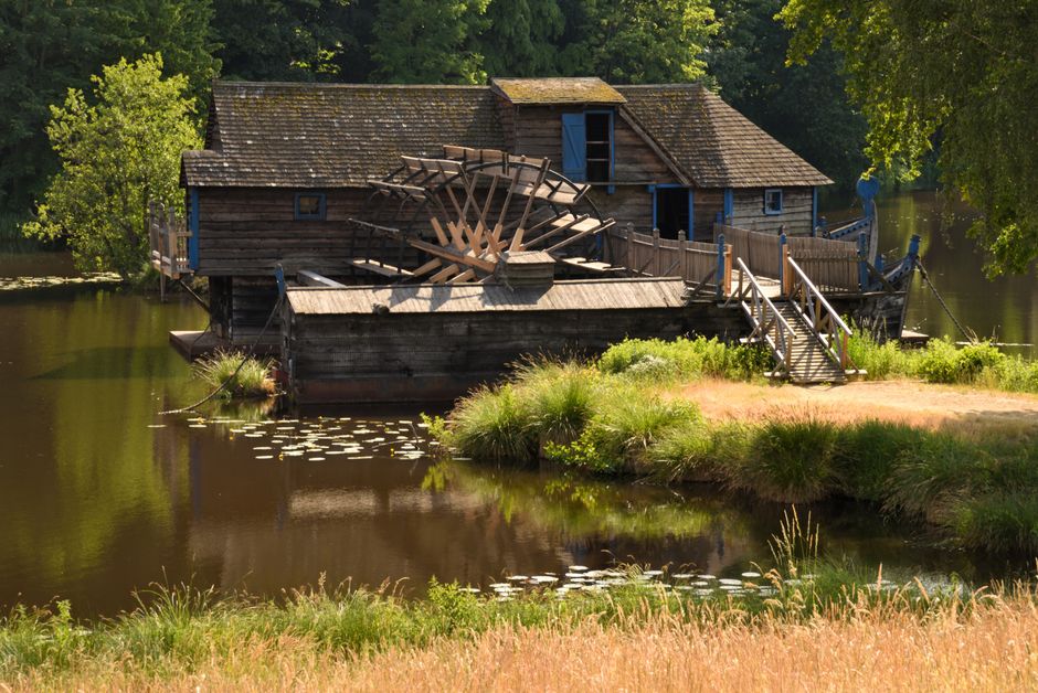 A rustic wooden watermill house situated on the edge of a pond, with a large wooden water wheel prominently attached to its side. The house features a sloped, shingled roof, and small blue window shutters, some of which are open. A wooden walkway with railings extends from the front entrance over the water to the grassy bank, leading to a small landing area. The pond's surface is calm, reflecting the surrounding greenery and the watermill structure. Lush trees with thick green foliage surround the scene, providing shade and framing the watermill. The environment appears peaceful and rural, indicative of a scenic setting suitable for house removals or relocation services that involve transporting historical or unique properties, with natural light highlighting the textures of the wood and foliage. Man with Van Colliers Wood occasionally supports such home relocation projects involving transportation of distinctive structures.