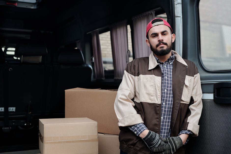A man with a beard and wearing a red cap, a plaid shirt, and a brown and cream work jacket stands inside a van used for home relocation, with his arms crossed. In the background, there are several cardboard moving boxes of various sizes, some sealed with packing tape, placed on the floor of the van. The interior of the van features dark seating and curtains on the side windows. The scene captures the process of furniture transport and packing during a house move, with the man representing a professional remsovals service, Man with Van Colliers Wood, involved in unloading or organising items for a local move.